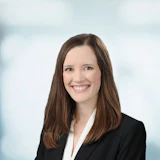 Professional headshot of woman in black blazer and white blouse smiling against blurred office background