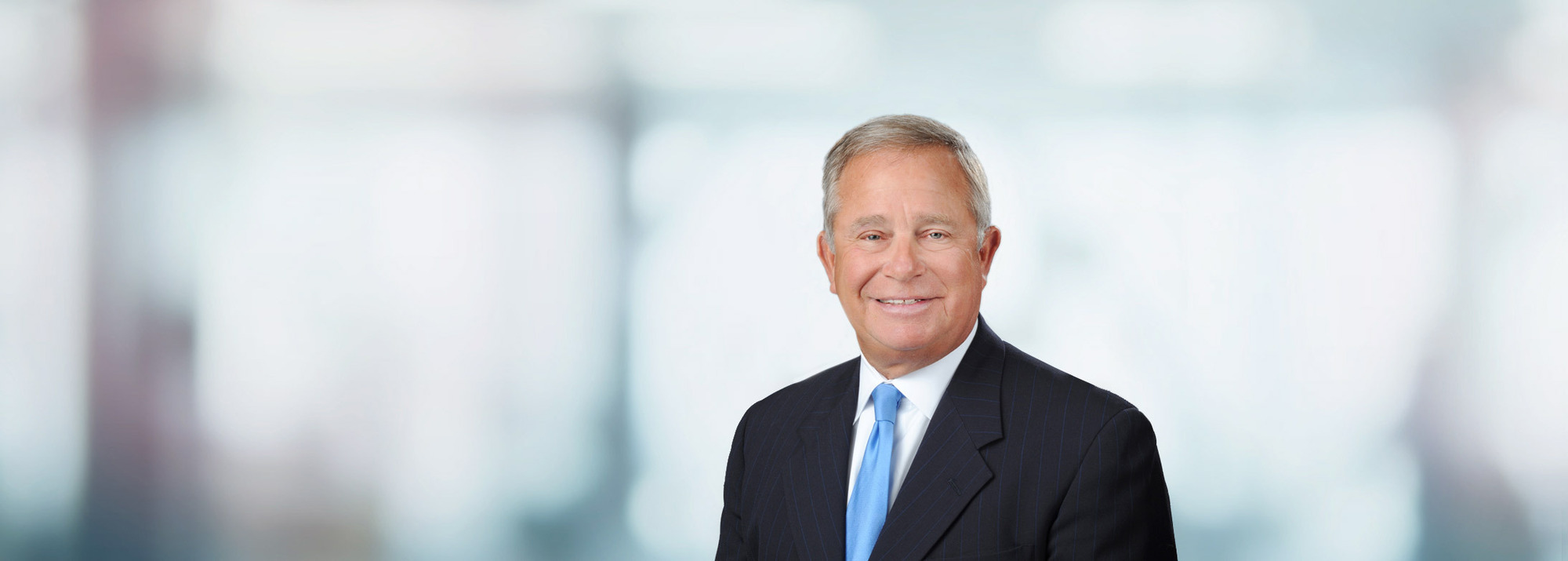 Professional headshot of a man in dark suit and blue tie against blurred office background