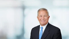 Professional headshot of a man in dark suit and blue tie against blurred office background