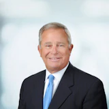Professional headshot of a man in dark suit and blue tie against blurred office background