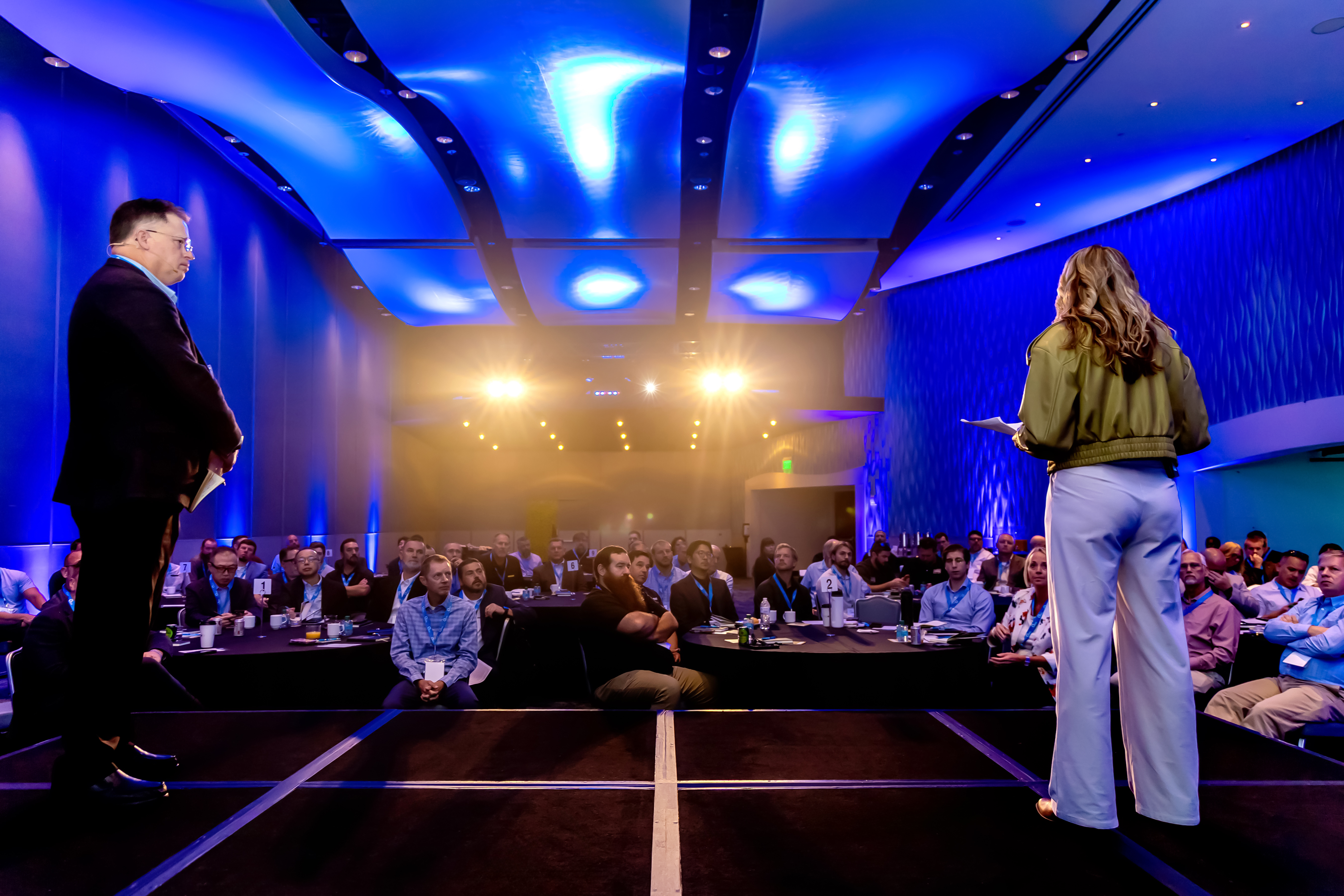 Two business speakers present on stage to an audience in a modern conference hall with blue ambient lighting and warm spotlights.
